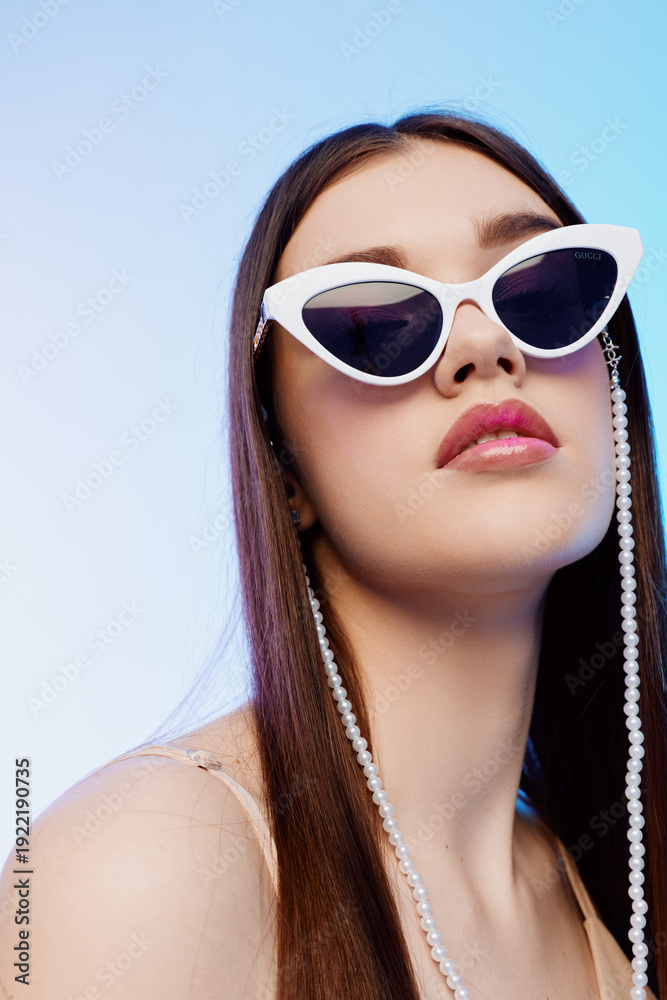 Naklejka premium Young woman beauty fashion wearing white sunglasses with pearl chain looks confident against blue background. Close-up portrait with long hair and pink lips. Stylish concept.