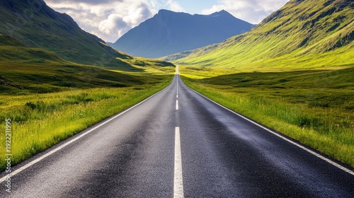 An endless asphalt road stretches through verdant green valleys towards sun-drenched mountains under a vast, cloudy sky.