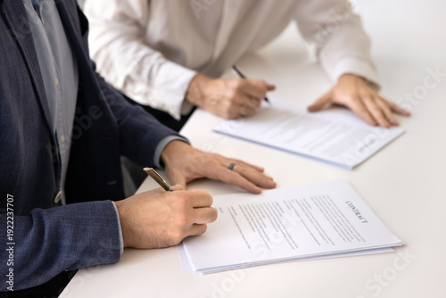 Formal commitment through contract signing. Close up view of two businessmen signing official documents seated at table. Legal validation, trust between parties and finalization of business agreement
