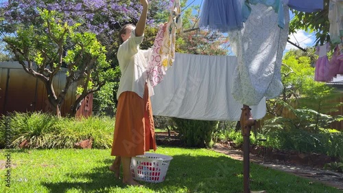 Young adult woman stands in her backyard and hangs clothes on a line while holding a basket of laundry on a sunny day.
