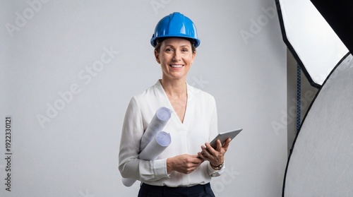 A smiling woman in a blue hard hat holds rolled plans and a digital tablet standing against a light background with studio equipment visible
