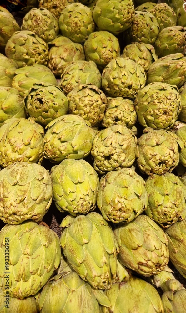 Obraz premium Fresh green artichokes are stacked closely together on a shelf, likely in a supermarket. The artichokes have a layered, scaly appearance with tightly packed leaves, displaying a bright green color