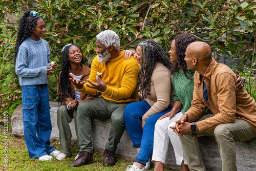 African American family with senior and child sitting on wooden log in garden, sharing white flower