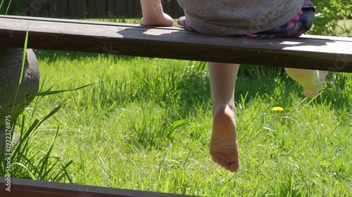 Fresh lawn. Child dangles his legs while sitting on wooden park bench, against backdrop of cheerful greens. The vibe is one of spring youth, mood of adolescence carefree bliss of childhood. 2026, 4K