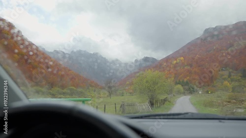 From the driver's seat, a narrow lane winds into a Balkan valley with orange and red autumn forest, a green gate, fence, rocky ridge, and low clouds in overcast light.