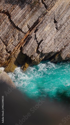 Aerial view of stratified limestone cliff in Montenegro as Adriatic waves crash into rocky inlets, frothy surf swirls, diagonal bedding and orange seams in bright sun