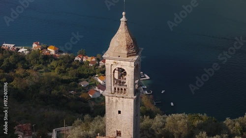 Aerial pan shows a weathered stone bell tower above olive groves and ruined roofs, terracotta houses, tiny piers, boats on a deep bay, and gentle wakes in warm light.