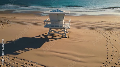 Lifeguard tower on sandy beach with clear blue ocean waves in the background, showcasing shadows and footprints on the sand during golden hour light