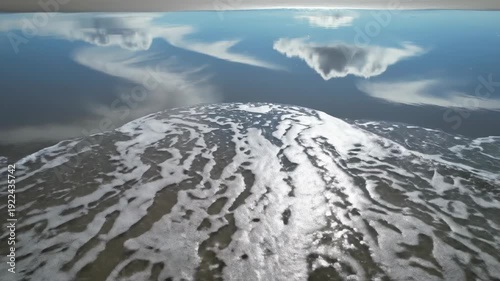 Calm ocean waves gently lap against the shore, reflecting clouds and sunlight, creating a serene coastal landscape with rippling water and foamy edges