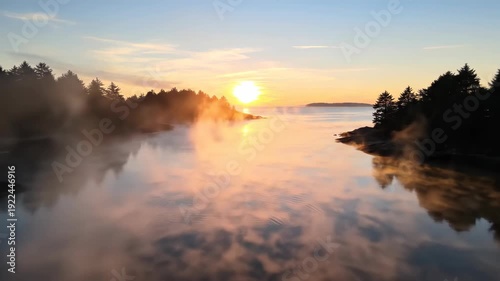 Sunrise over tranquil water with mist rising from the surface, surrounded by evergreen trees and rocky shoreline, creating a serene natural landscape