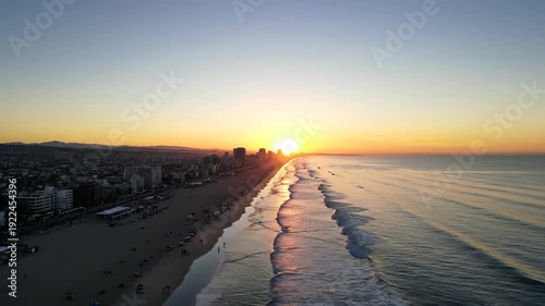 Aerial view of sunset over beach with gentle waves lapping at shoreline, city skyline visible in background, creating a serene coastal atmosphere