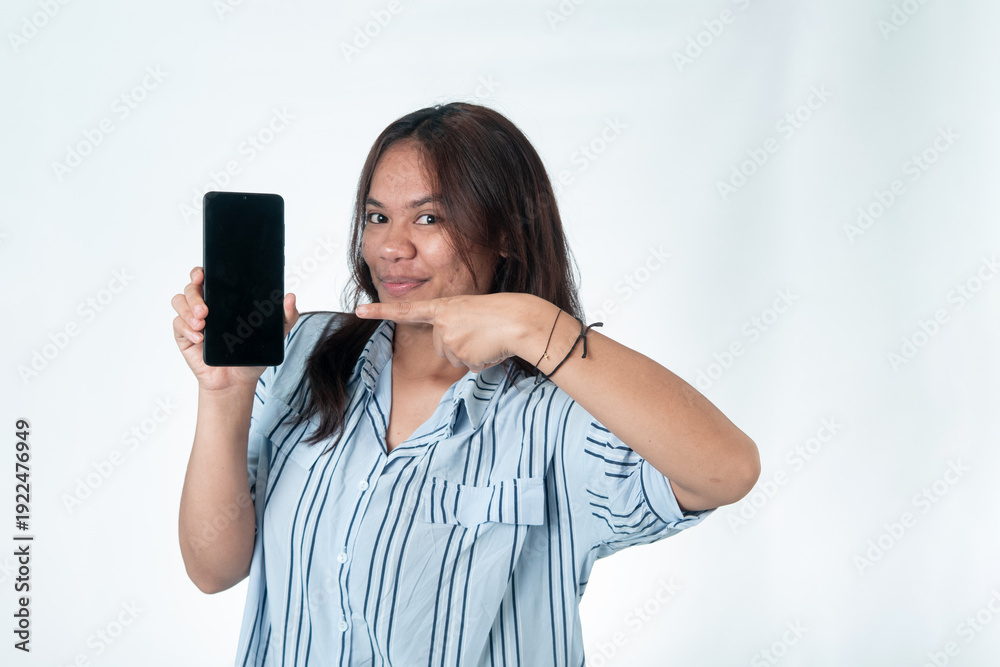 Fototapeta premium Indonesian woman in a blue striped shirt holding up a smartphone and pointing her finger at the black screen. Smiling expression, looking at the camera. Isolated on white. 