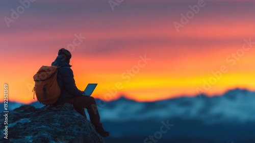 Summer for digital nomads, A person with a backpack sits on a rock, working on a laptop against a vibrant sunset over mountainous terrain.