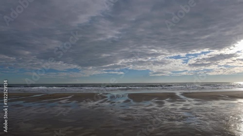 Serene coastal landscape showing gradual wave movement and shifting cloud patterns over sandy beach with reflections on wet surface during daylight hours