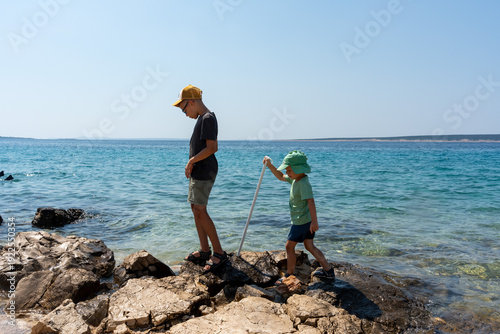 Two boys brothers in caps exploring rocky adriatic coastline together with stick on hot sunny summer day with turquoise sea background. Sibling bonding, childhood adventure and family vacation.