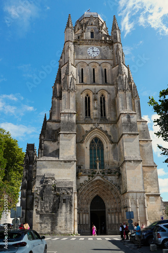 Main facade and ornate bell tower of the Basilica of Saint-Eutrope in Saintes, France. The historic Romanesque and Gothic architecture features a large clock and intricate stone carvings under a blue 