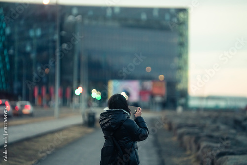 Bilde på lerret Woman in black coat filming a video with smartphone at Reykjavik waterfront