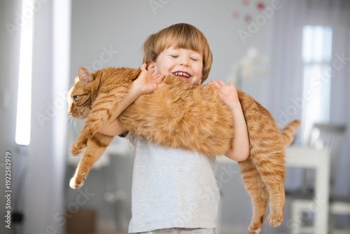 Toddler boy holding ginger cat on hands at home with light interior. Cute funny picture of kids and pets friendships. Life with domestic cat. Fluffy ginger tabby cat. Pets life.