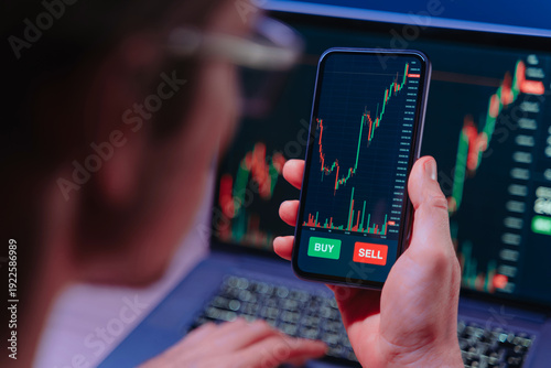 Over shoulder view of crypto trader analyzing live crypto and stock charts on smartphone with trading platform app, displayed on laptop and phone screen in dark neon lit room.