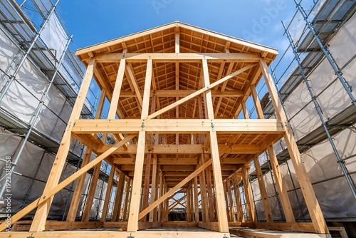 Obraz na płótnie 青空の下で建築が進む木造住宅の構造躯体と周囲に設置された建設現場の足場とメッシュシート