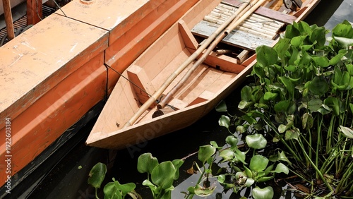 A traditional wooden sampan boat floating next to an orange industrial hull in water filled with green water hyacinths. High-angle view showcasing manual and industrial river maintenance.