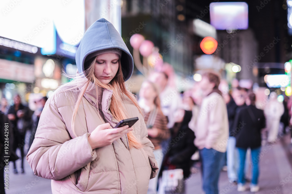 Fototapeta premium Teenage girl using smartphone at Times Square New York at night, young woman in winter hooded jacket standing among crowd and neon lights in busy urban street