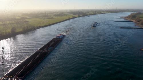 Wallpaper Mural Aerial view of a long industrial barge transporting bulk cargo down a wide river at sunset. Wind turbines and misty green fields line the horizon under a soft golden glow. Torontodigital.ca