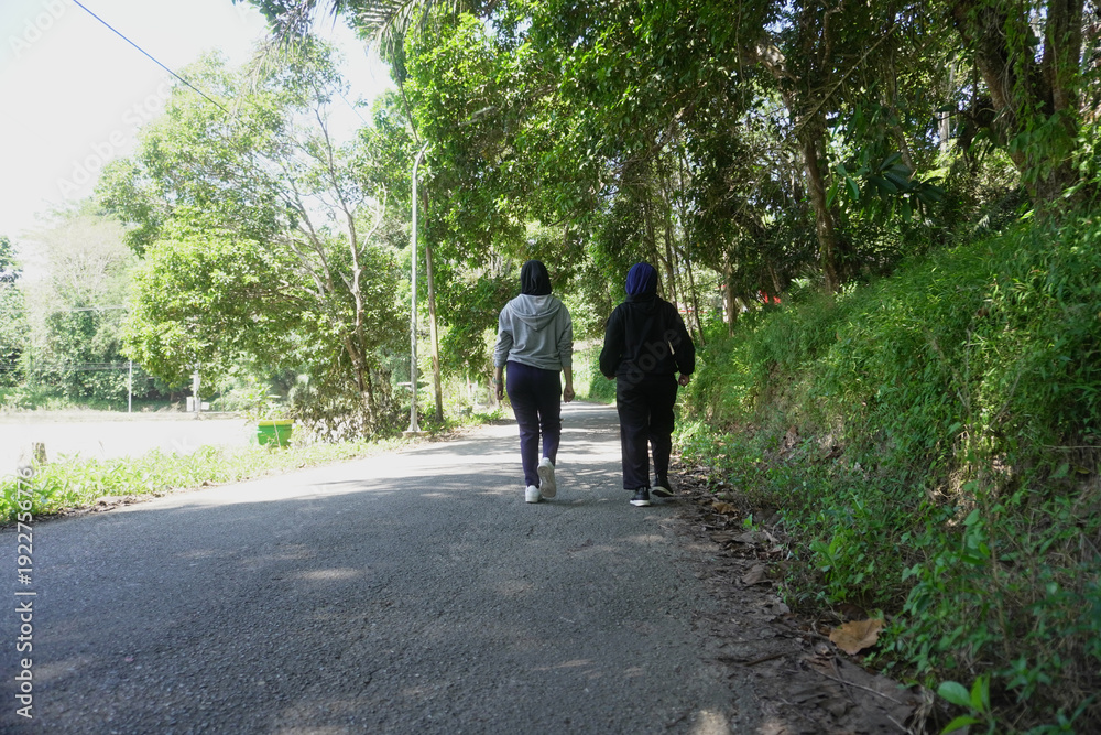 Fototapeta premium Two young Muslim women walking together for morning exercise along a green park pathway filled with trees on a bright sunny day. 