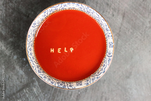 Tomato soup in a blue bowl with a word Help in it made of vermicelli. Grey textured background with space for text. 