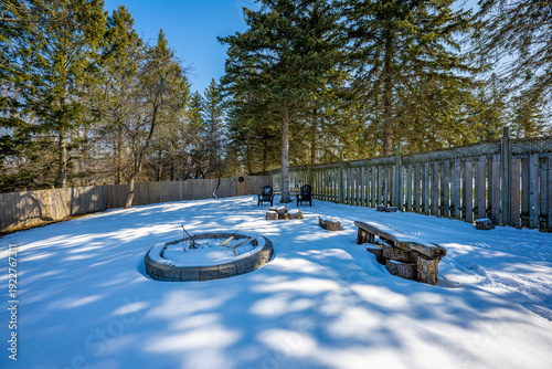 Backyard in ramara, ontario, featuring a circular stone fire pit, adirondack chairs, a rustic wooden bench, and pine trees, all covered in a blanket of fresh winter snow on a sunny day