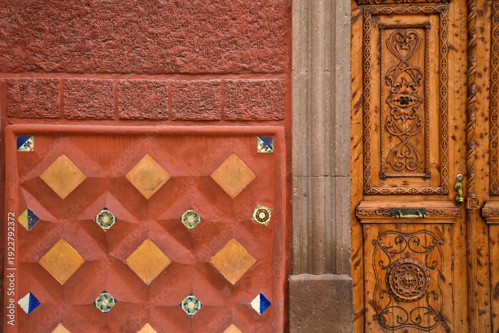 Fototapeta premium Facade view of a Colonial architecture house with a Venetian red stucco wall, a hand carved wooden door and a terracotta ceramic tile decoration, in San Miguel de Allende, Guanajuato Mexico.