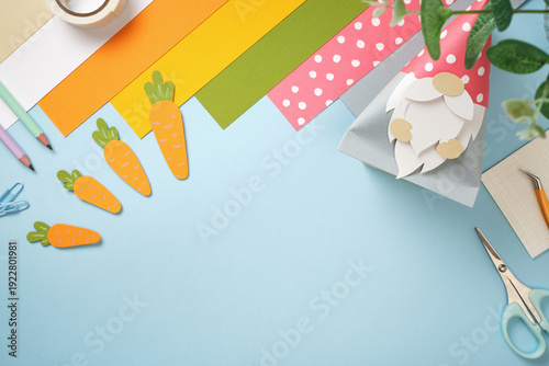 Easter paper crafts with a gnome bunny, carrots, and colorful stationery on a blue background. Spring DIY project. Copyspace.