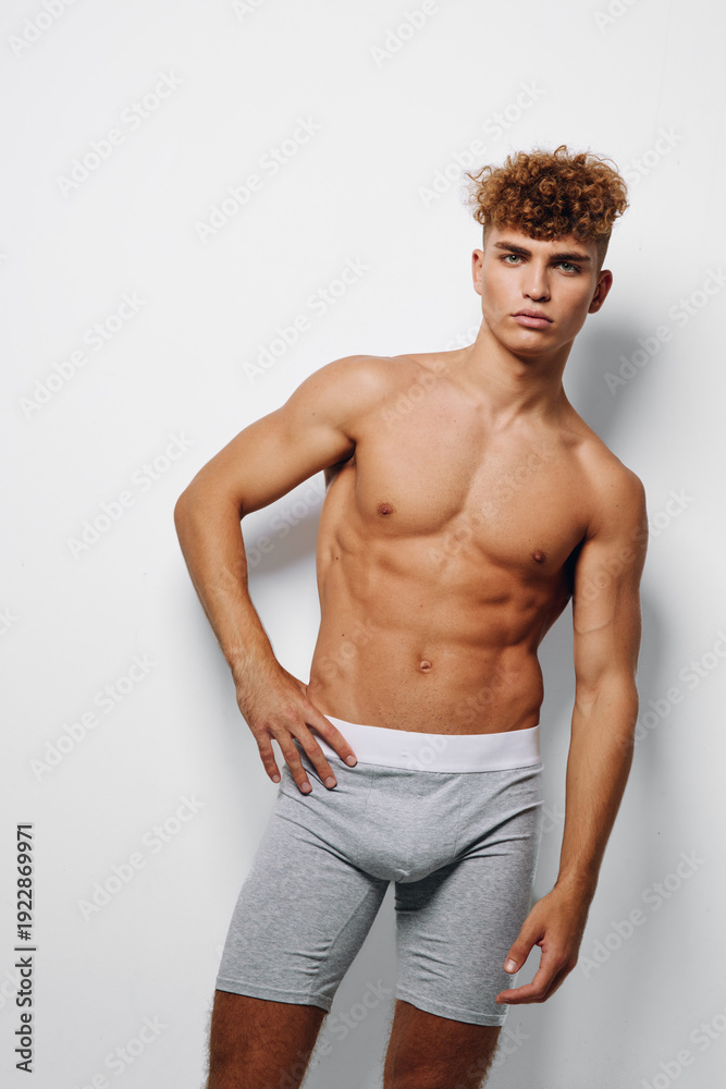 Fototapeta premium Male model standing shirtless against white wall wearing gray shorts with fitted neckline for modern fitness fashion and athletic body showcasing toned muscles and confident pose with minimal