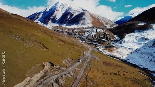 Drone view of car is driving along valley approach with scattered stone houses and snowy peaks, sweeping slopes bathed in soft light, river path leading into historic village, sense of remote travel
