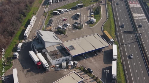 Highway Truck Stop: Aerial perspective captures a bustling highway truck stop, where numerous trucks are parked for rest. alongside a flowing highway.