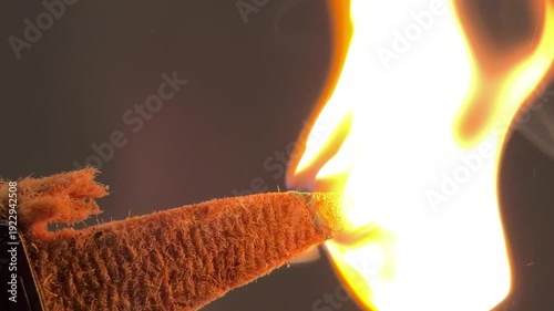 A hand holds a specialized tourist match with a thick brown body and green tip. The match is being pulled across the textured striking surface of a matchbox in a close-up indoor shot.