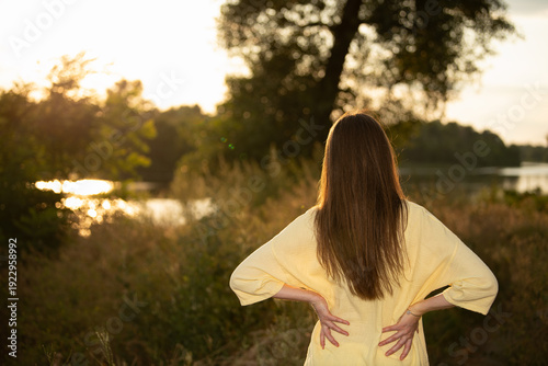 A woman stands by a lake with her hands on her hips. She wears a yellow shirt and looks at the water as the sun sets. Trees surround her