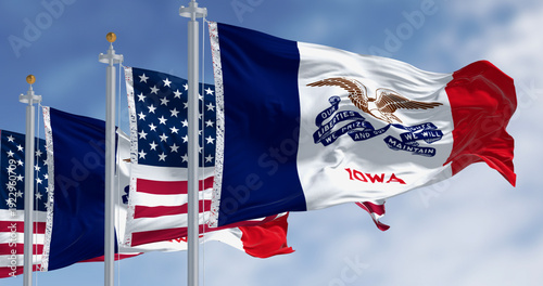 Detailed 3D digital illustration of the Iowa state flag and American national flag waving against a clear blue sky. Realistic fabric textures and lighting on dual flagpoles.