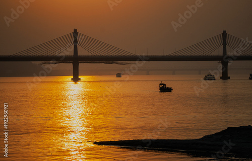 Silhouetted boats move across the Brahmaputra River at sunset beneath a cable-stayed bridge, as golden light reflects across the calm water