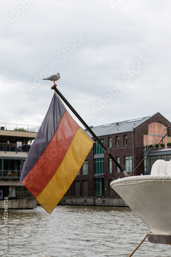 A seagull perched on a flagpole with the German national flag on the stern of a boat in the harbor, with city buildings and water in the background.