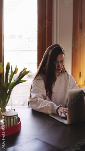 Focused young woman working on laptop at home office, serious girl typing on keyboard and then smiling, remote work and freelancer lifestyle concept, cozy interior with yellow tulips
