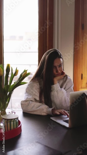 Tired young woman working on laptop at home office, bored or pensive girl sitting at desk with yellow tulips, burnout and work from home stress concept
