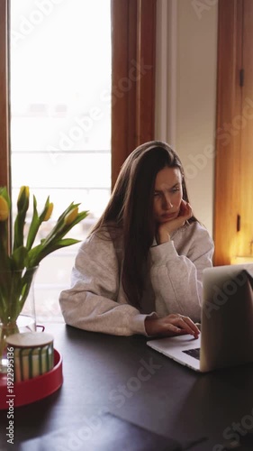 Tired young woman working on laptop at home office, bored or pensive girl sitting at desk with yellow tulips, burnout and work from home stress concept