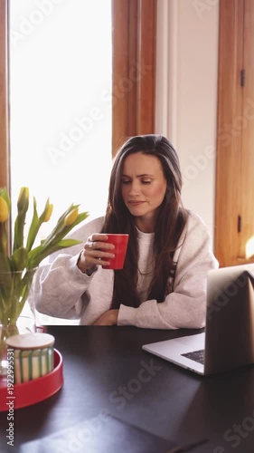 Young woman working on laptop and drinking from red cup at home office, happy freelancer taking a sip of coffee or tea during work, cozy workspace with yellow tulips
