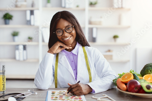 A nutritionist sits at a table with a notebook, pen in hand. Fresh fruits and vegetables are nearby. She smiles while assessing meals. A glass of juice is also visible in the setting.