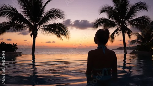 Silhouette of a woman relaxing in an infinity pool overlooking the ocean at sunset with tropical palm trees