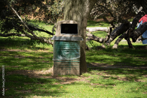 Obraz na plátně Park trash can with plastic liner sits beneath a large tree with dappled sunligh