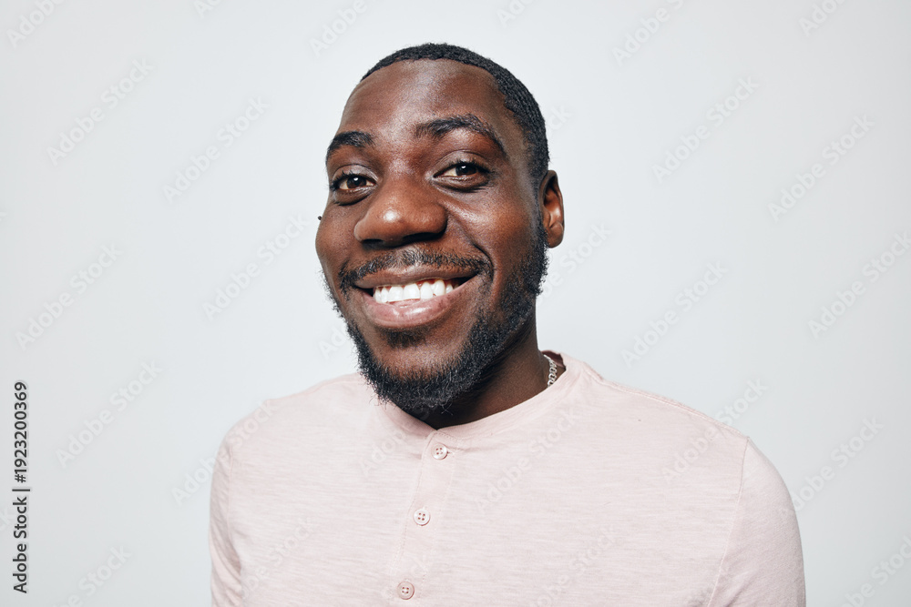 Obraz premium African American man with dark skin smiling and looking at camera in casual light pink shirt. Portrait of happy black male with beard and short hair on white background.