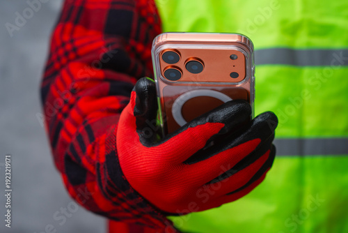 Φωτογραφία Man worker wearing a high-visibility vest and protective gloves uses a mobile phone at a building site