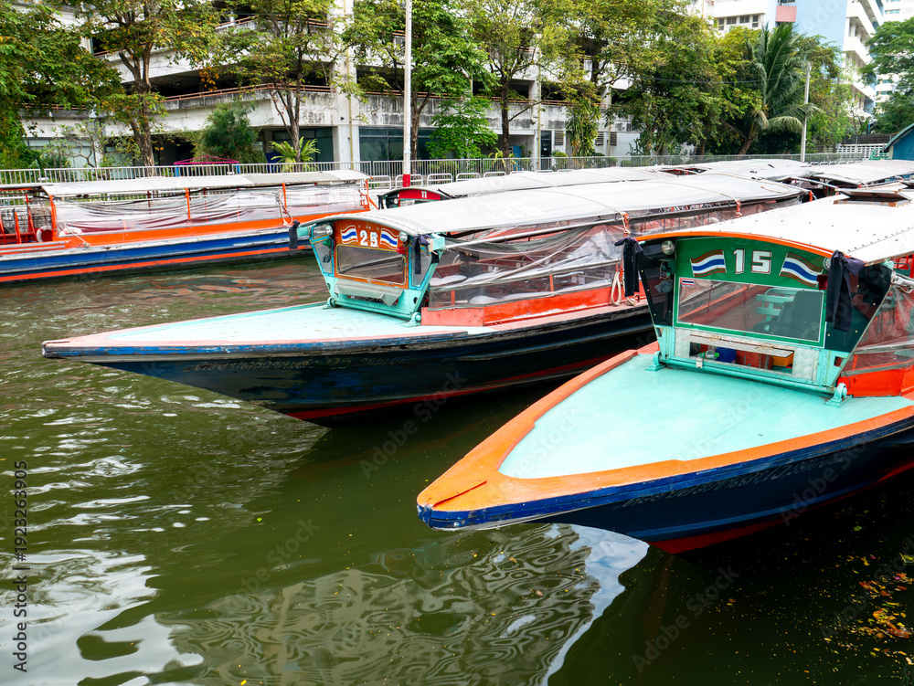 Obraz premium Public passenger boats running on canal in Bangkok city with buildings and trees on bank, water transportation and urban commuting travel.